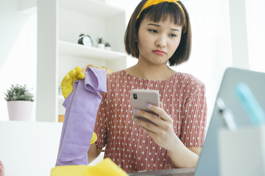 亚洲女佣在起居室清洁家具，工作时使用智能手机。女佣服务申请。