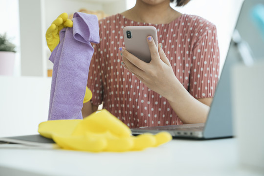 亚洲女佣在起居室清洁家具，工作时使用智能手机。女佣服务申请。