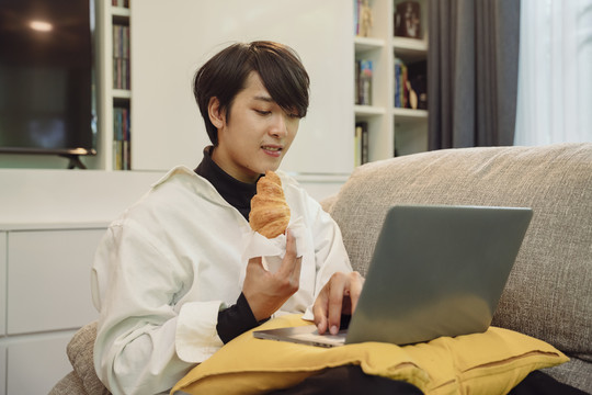 商人在家工作，一边用笔记本电脑吃新鲜烤羊角面包。