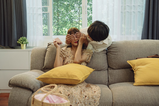 当女人坐在沙发上时，亚洲男人惊讶地从背后遮住了她的眼睛。家里有一对顽皮的夫妇。