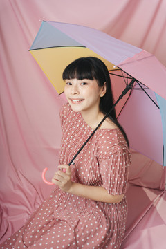 美丽的泰国亚裔长发女子身穿粉色连衣裙，手持彩虹伞，背景为粉色。时尚肖像。