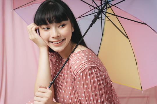 美丽的泰国亚裔长发女子身穿粉色连衣裙，手持彩虹伞，背景为粉色。时尚肖像。