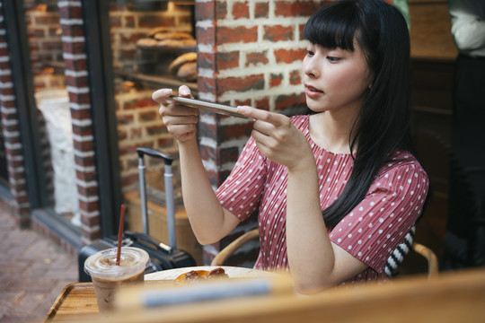 美丽的亚泰长黑发女子在咖啡厅用智能手机拍面包蛋糕。先吃饭。