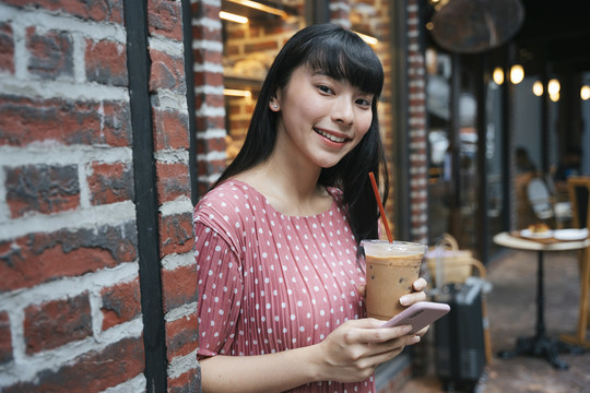 曼谷街头，一位美丽的亚泰黑发长发女士，手拿冰咖啡和智能手机。