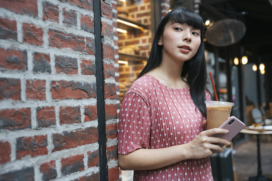 曼谷街头，一位美丽的亚泰黑发长发女士，手拿冰咖啡和智能手机。