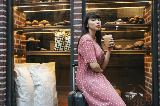 在面包店橱窗前的街道上，一位美丽的亚泰裔黑发女子拿着吸管喝冰咖啡。