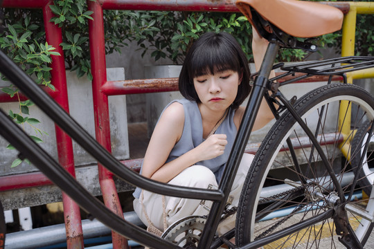 一位年轻的泰国亚裔女子坐在一辆坏了的自行车旁，沮丧地等着别人来修理。