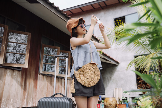 美丽的亚泰女旅行家，穿着夏季休闲服，戴着帽子，带着当地酒店的手推车行李。本地旅行。