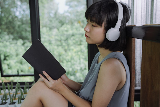 侧视图-美丽的亚泰女人深色短发，戴着耳机听音乐，在当地的旅馆房间看书。