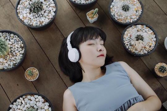 俯瞰图-美丽的亚泰女人深色短发闭着眼睛躺在木地板上用无线耳机听音乐。