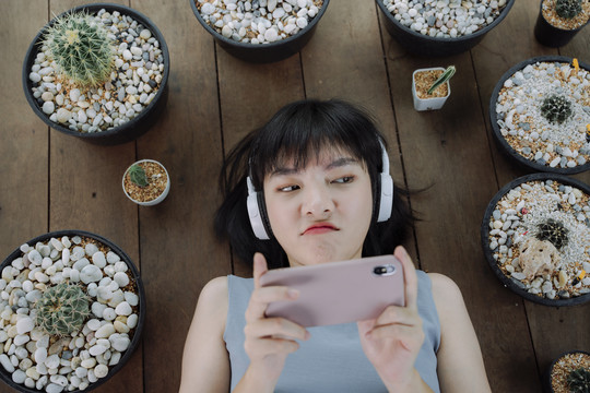 顶视图-美丽的亚泰女性短发，戴着耳机，在智能手机上玩游戏。