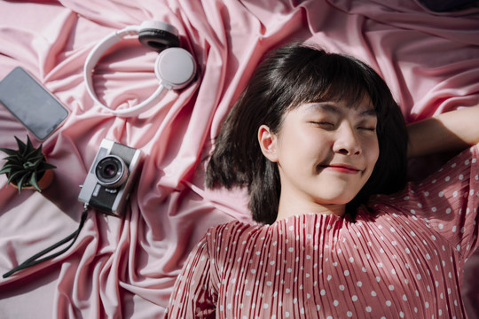顶视图-美丽的亚泰女人黑色短发睡在粉红色背景。相机，耳机，智能手机。旅行梦想家。