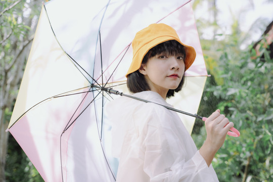 时尚写真——美丽的亚泰女人深色短发，穿着白色外套，戴黄色帽子，在雨季撑着柔和的彩虹伞。