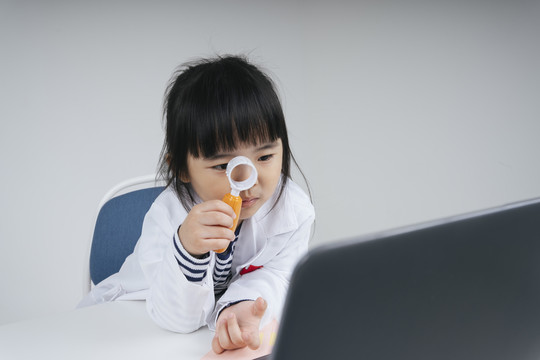 漂亮的泰国亚裔小孩扮演医生的角色拿着放大镜看笔记本电脑。