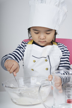 漂亮的泰国亚裔小孩扮演厨师的角色，在厨房的玻璃碗里混合面粉和水。