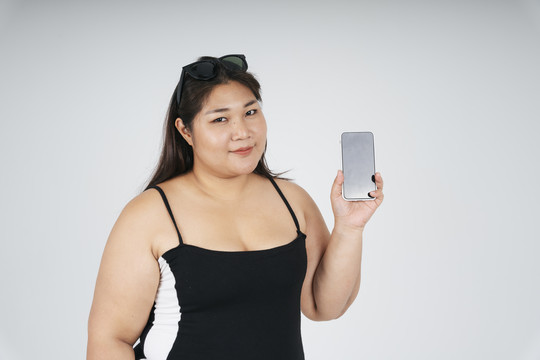 身着黑色连衣裙的亚洲胖女人的肖像，在白色背景上显示智能手机屏幕，供模拟或复制空间使用。