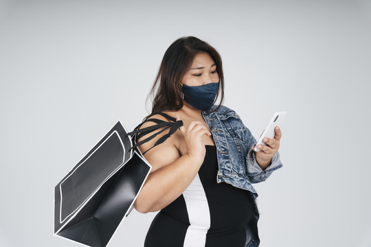 健康的亚洲胖女人拿着购物袋在白色背景上用智能手机在线订购的肖像。