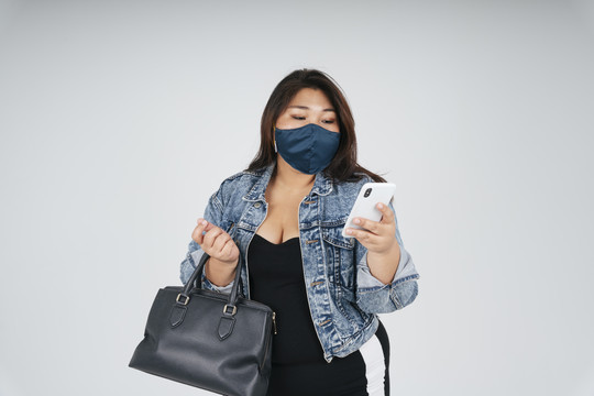 健康的亚洲胖女人戴着面具，穿着牛仔裤夹克，在白色背景下使用智能手机进行网上购物。