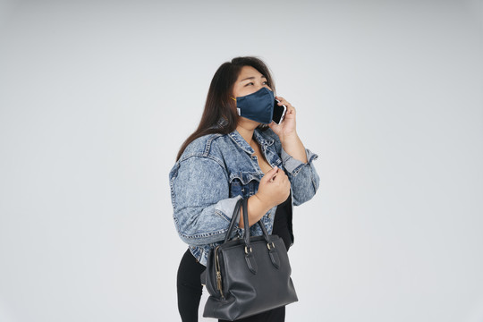 健康的亚洲胖乎乎的女人戴着面具，穿着牛仔裤夹克，在白色背景下打电话。