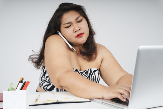 忙碌的亚洲胖乎乎的女人拿着笔记本电脑和电话，严肃地看着白色背景。
