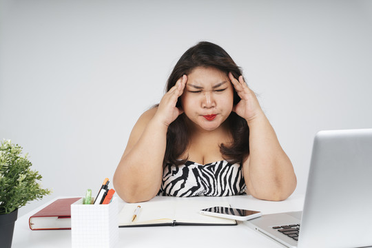 紧张的亚洲胖女人坐在办公桌前工作时头痛。