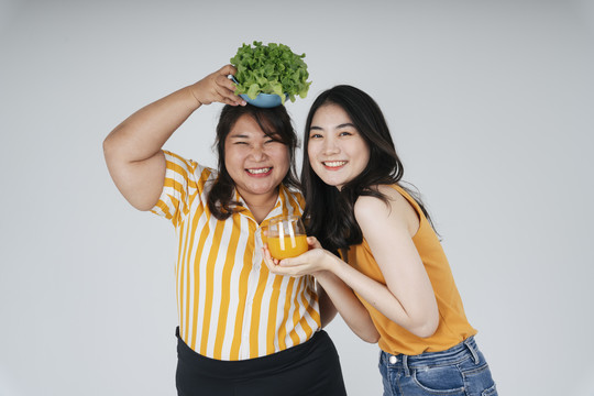 两位亚洲健康女性，白色背景，配沙拉碗和橙汁。
