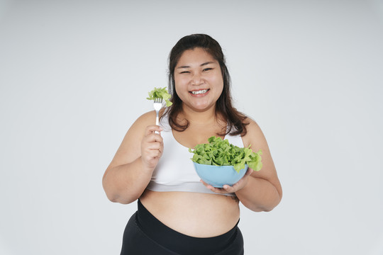 快乐快乐的亚洲胖女人穿着白色运动胸罩，喜欢吃白色背景的沙拉碗。