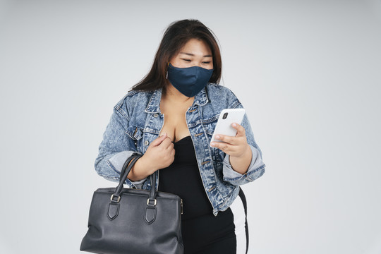 购物情人的画像穿着黑色连衣裙和牛仔裤夹克的胖女人戴着面具在智能手机上使用在线购物应用程序感到兴奋。