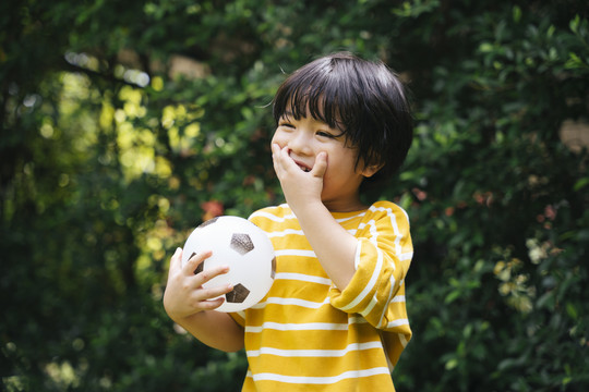 快乐的亚洲小孩拿着一个足球盖嘴，手在公园里笑着。