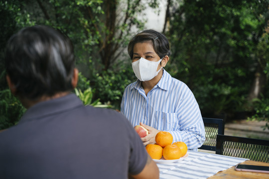戴着口罩持刀剥水果的亚洲老太太在公园与丈夫在户外交谈。