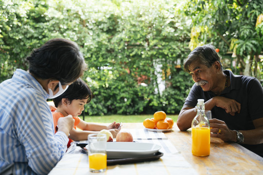 爷爷奶奶和孙子在院子里野餐。有趣的爷爷看着一个男孩在智能手机上玩游戏。