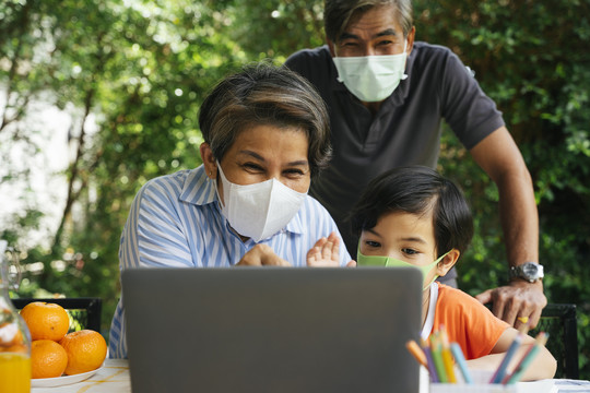 亚洲人的祖父母和孙子戴着口罩在院子里和一个坐在笔记本电脑上的人打视频电话。