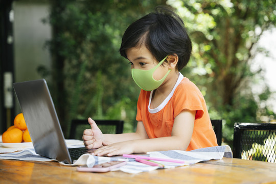 侧视图-亚洲小孩戴着口罩在院子里用笔记本电脑。在家学习。