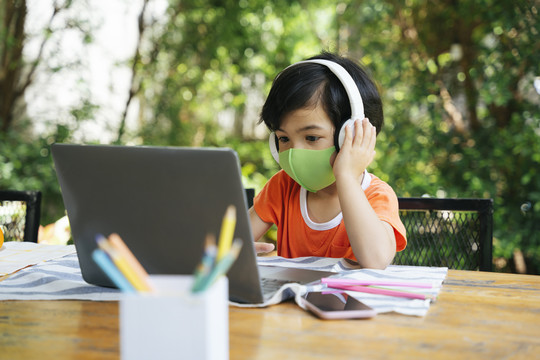 亚洲小孩戴着口罩在院子里用耳机和笔记本电脑听音乐。