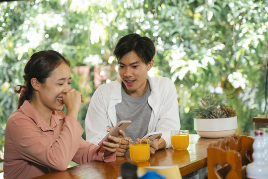 一对亚洲年轻夫妇用智能手机在社交媒体上看东西。两人都坐在木条上喝橙汁。