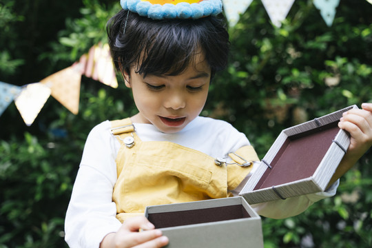 快乐的亚洲黑发小孩戴派对帽在院子里拿着礼物盒。