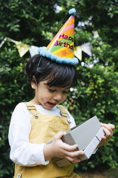 快乐的亚洲黑发小孩戴派对帽在院子里拿着礼物盒。