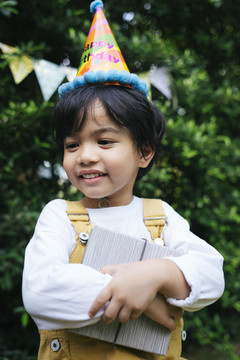 快乐的亚洲黑发孩子戴着派对帽，手里拿着礼盒在院子里。