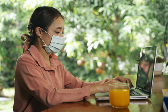 侧视图-戴着面具的亚裔妇女在吟游诗人的笔记本电脑上工作，喝着一杯橙汁。