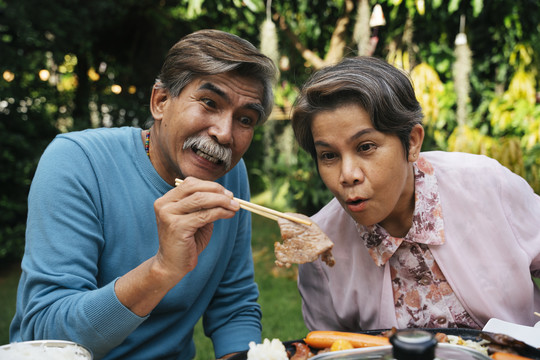 惊讶的亚洲长辈夫妇用筷子从电子锅里拿起烤肉。两人都惊讶地看着猪肉，神情激动。