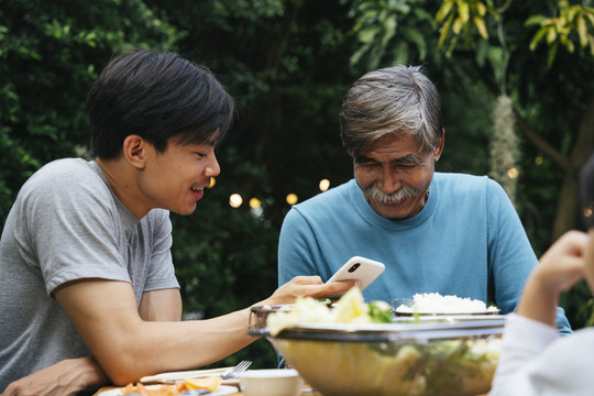 亚裔儿子在户外的饭局上用智能手机给父亲看东西。