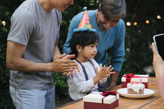 亚洲小孩在户外和家人一起庆祝生日聚会。妈妈用智能手机拍照。