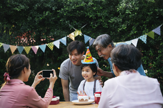 亚洲小孩在户外和家人一起庆祝生日聚会。妈妈用智能手机拍照。