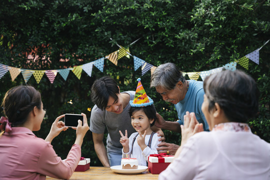 亚洲小孩在户外和家人一起庆祝生日聚会。妈妈用智能手机拍照。