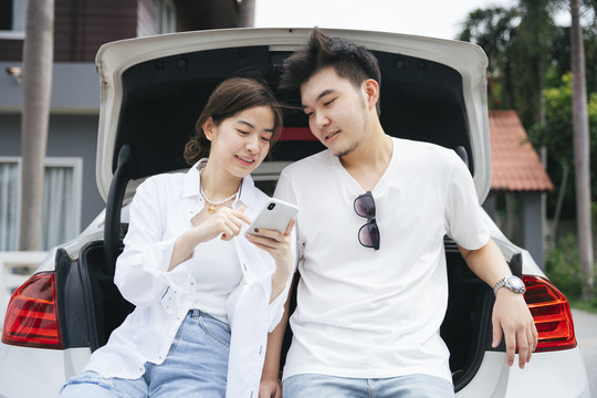一对亚洲夫妇坐在汽车后备箱旁，通过智能手机应用程序查看目的地。
