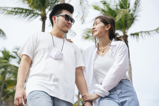一对穿着白衬衫的亚洲情侣手牵着手在沙滩上散步。