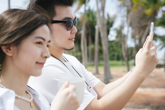 戴着墨镜的亚洲人拿着智能手机拍着大海的照片，和妻子站在沙滩上。