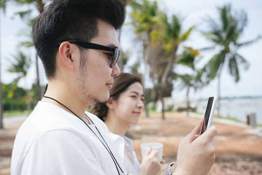 戴着墨镜的亚洲人拿着智能手机拍着大海的照片，和妻子站在沙滩上。