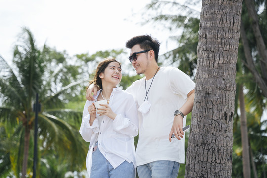 一对亚洲情侣肩并肩站在椰树附近的海滩上。