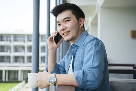 穿着蓝领衬衫的亚洲商人在酒店阳台上打电话。